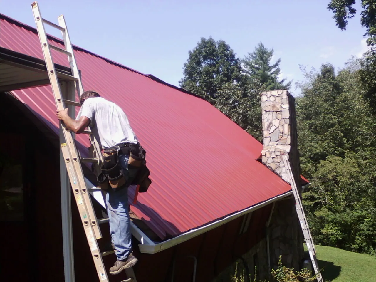 Roofer on a ladder beside a red metal roof and stone chimney — Soffit & Fascia Repair by Roofing Brothers in Commerce City